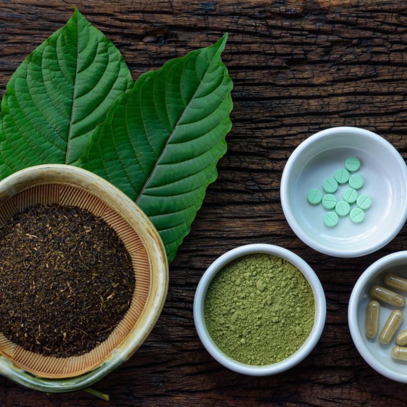 Mitragyna speciosa (kratom) leaves with medicine products in powder, capsules and tablet in white ceramic bowl with wooden texture on background